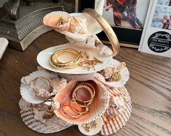May include: A tiered jewelry organizer constructed from various natural seashells, featuring a central white ceramic dish. The structure holds an assortment of gold-toned metal rings, hoop earrings, a thick textured bangle, and delicate chain necklaces.