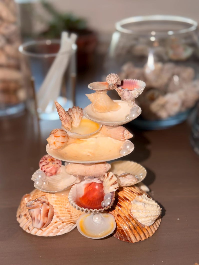 May include: A tiered decorative sculpture constructed from various seashells and small white faux pearls. The structure features multiple levels of stacked bivalve and spiral shells in shades of cream, tan, orange, and brown, arranged in a conical shape.