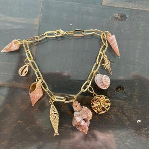 May include: Gold-toned charm bracelet featuring various seashell and marine life charms. The bracelet includes a fish, starfish, and a round charm with a sunburst design. The chain is made of large oval links, and the charms are in shades of tan and gold.