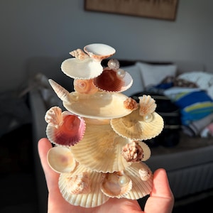 May include: A decorative sculpture constructed from various seashells arranged in a tiered, vertical formation. The piece features a mix of scallop, spiral, and bivalve shells in shades of cream, tan, and pink, accented with small white spherical pearls.