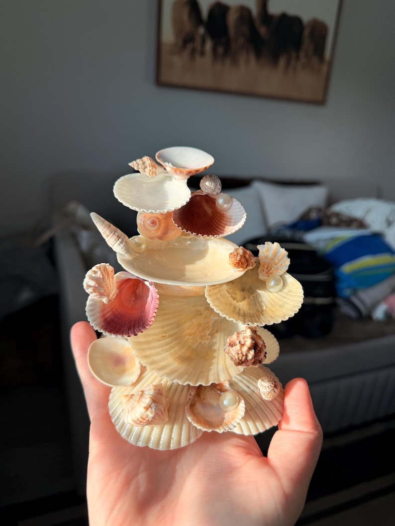 May include: A decorative sculpture constructed from various seashells arranged in a tiered, vertical formation. The piece features a mix of scallop, spiral, and bivalve shells in shades of cream, tan, and pink, accented with small white spherical pearls.