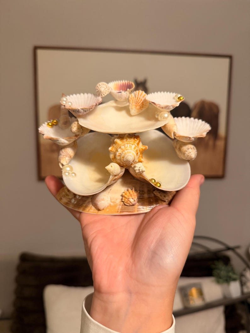 May include: A tiered decorative display piece constructed from various natural seashells. The structure features multiple white and tan scallop shells arranged in layers, accented with small white pearls and gold beads, centered around a spiral shell.