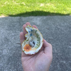 May include: A decorative oyster shell featuring a hand-painted interior with a floral motif including blue flowers, a green fern, and an orange flower. The shell has a gold-painted scalloped edge and a small, dark, textured central accent.