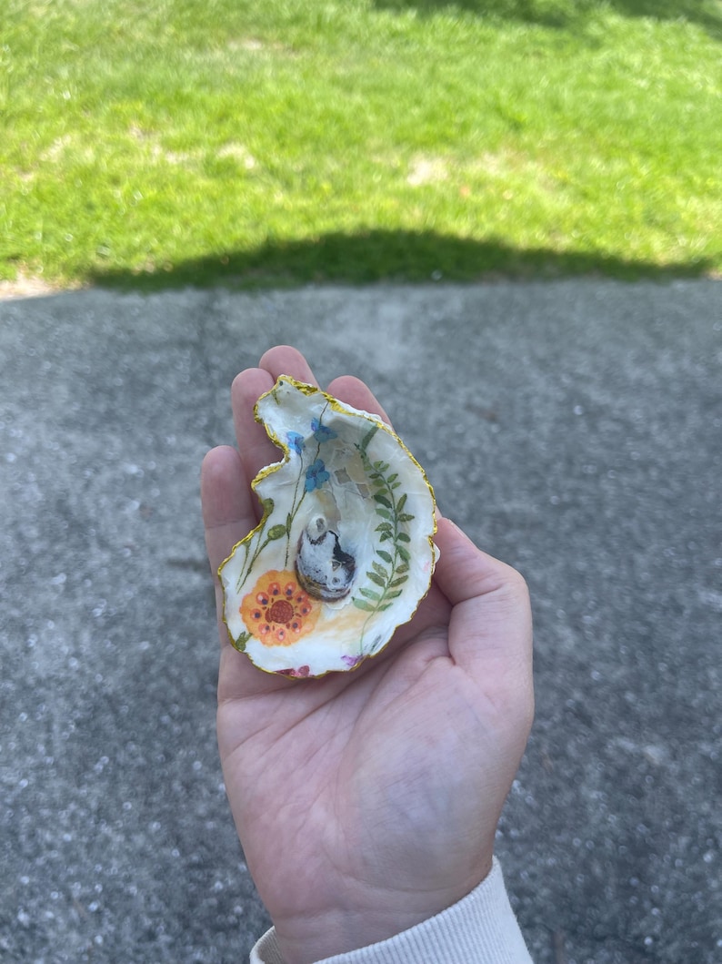 May include: A decorative oyster shell featuring a hand-painted interior with a floral motif including blue flowers, a green fern, and an orange flower. The shell has a gold-painted scalloped edge and a small, dark, textured central accent.