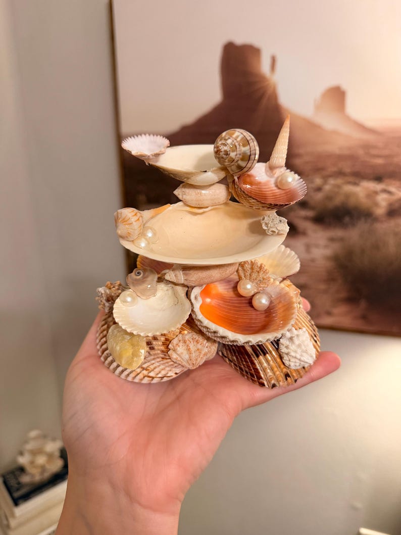 May include: A tiered decorative sculpture constructed from various seashells, including scallop, spiral, and clam shells. The structure features multiple levels with small white pearls nestled within the shells, showcasing natural tan, orange, and white tones.