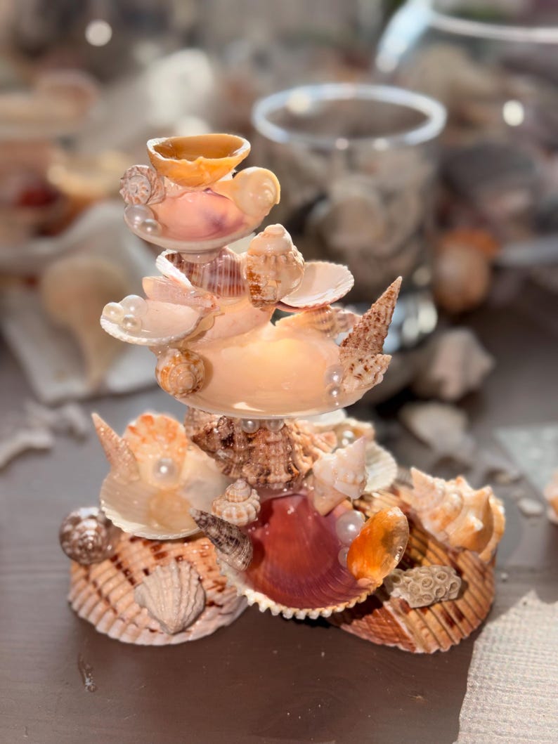 May include: A tiered decorative sculpture constructed from various seashells in shades of tan, brown, and cream, accented with small white spherical beads. The structure features multiple layers of shells arranged in a vertical, stacked formation.