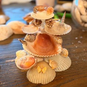 May include: A tiered decorative sculpture constructed from various seashells, including scallop, spiral, and conch shells. The structure features a central large shell base, smaller shells arranged in layers, and small white pearl accents throughout the design.