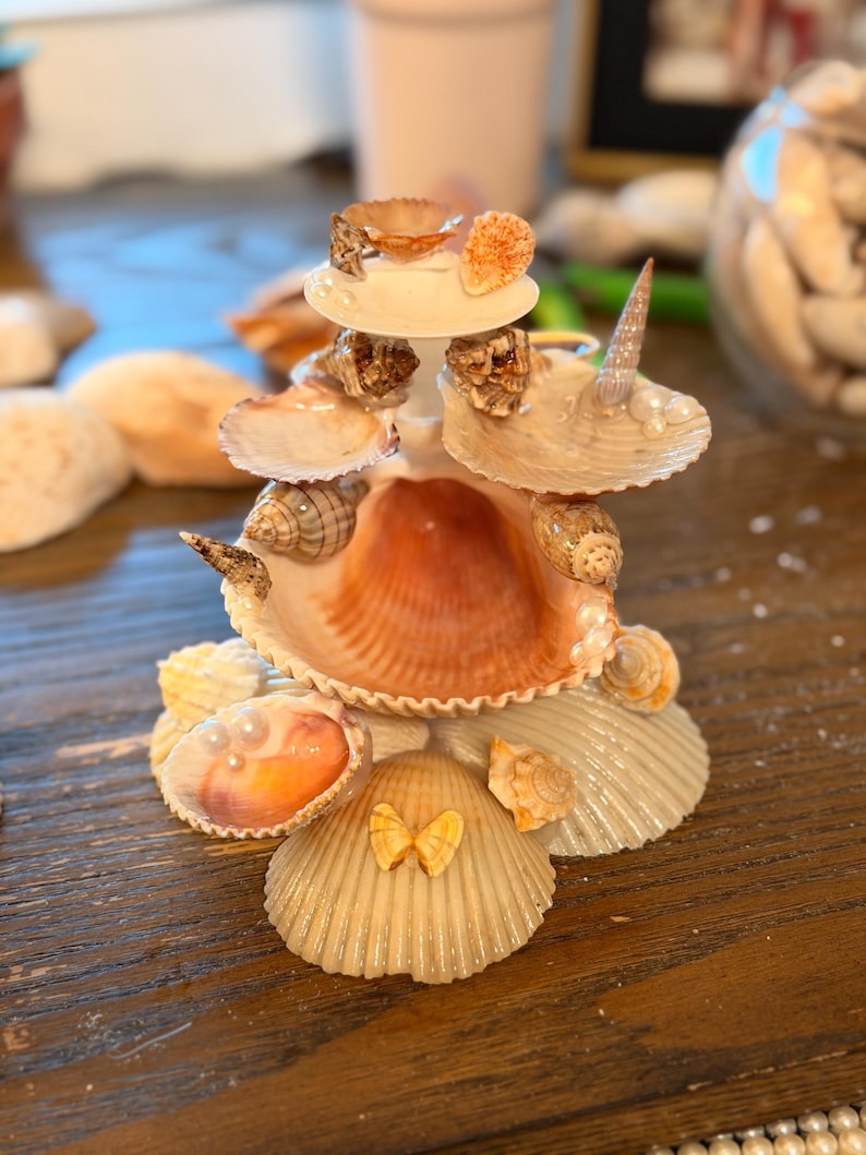 May include: A tiered decorative sculpture constructed from various seashells, including scallop, spiral, and conch shells. The structure features a central large shell base, smaller shells arranged in layers, and small white pearl accents throughout the design.
