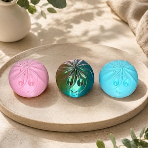 May include: Three spherical fidget toys with a swirled, dumpling-like design and a smiling face. The left toy is matte pink, the centre toy has a metallic iridescent finish shifting between green and blue, and the right toy is a solid matte light blue.