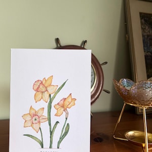Könnte beinhalten: Ein Aquarellgemälde von drei Narzissen in Gelb- und Orangetönen mit grünen Stielen und Blättern auf einer weißen Karte. Das Wort "DAFFODIL" ist unten in Schwarz geschrieben. Das Gemälde wird auf einer Holzoberfläche ausgestellt.