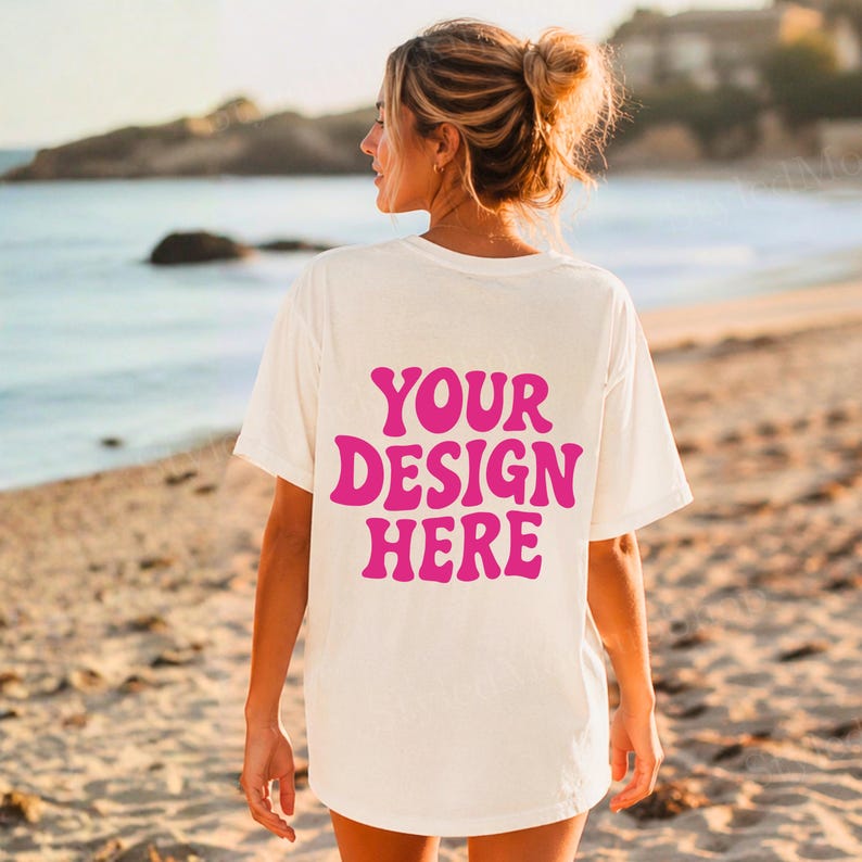 Puede incluir: Camiseta color crema con el texto "YOUR DESIGN HERE" en rosa brillante en la espalda. La camiseta la lleva una persona en una playa, con el mar y la arena de fondo.