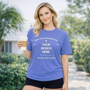 May include: A woman wearing a periwinkle blue t-shirt with the text "YOUR DESIGN HERE". The shirt is paired with black shorts. She is holding a white coffee cup with a brown sleeve. The background features greenery.