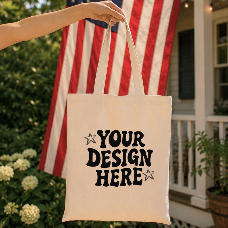 Puede incluir: Bolso tote de color crema con texto negro "YOUR DESIGN HERE" con detalles de estrellas. El bolso se sostiene frente a una bandera estadounidense. El bolso tiene dos asas y est&aacute; hecho de una tela resistente.
