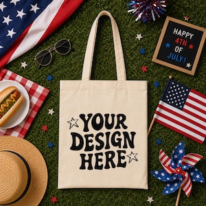 Puede incluir: Un bolso tote crema con el texto "YOUR DESIGN HERE" en negro, rodeado de estrellas. La imagen incluye un perrito caliente, gafas de sol, un sombrero de paja, una bandera americana y un letrero que dice "HAPPY 4TH OF JULY!"
