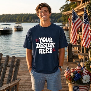 May include: Navy blue t-shirt with the text "YOUR DESIGN HERE" in white, accented with red stars. The shirt is worn by a person standing on a wooden dock, with American flags and boats in the background.