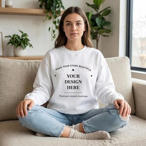 May include: A white sweatshirt with the text "MAKE YOUR STORE BEAUTIFUL YOUR DESIGN HERE" in black. The sweatshirt is a mockup, perfect for showcasing designs. The model is wearing light blue jeans and sitting on a beige couch.