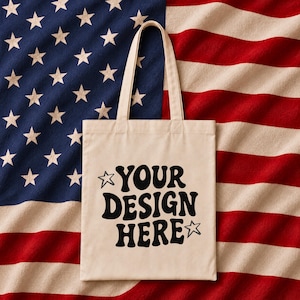 Puede incluir: Una bolsa de mano color crema con el texto "YOUR DESIGN HERE" en negro, sobre un fondo de la bandera estadounidense. La bolsa tiene dos asas y un dise&ntilde;o sencillo.