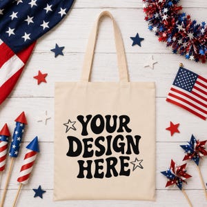 Puede incluir: Un bolso tote color crema con el texto "YOUR DESIGN HERE" en negro. La imagen presenta decoraciones patri&oacute;ticas, incluyendo una peque&ntilde;a bandera americana, adornos en forma de estrella y fuegos artificiales rojos, blancos y azules.