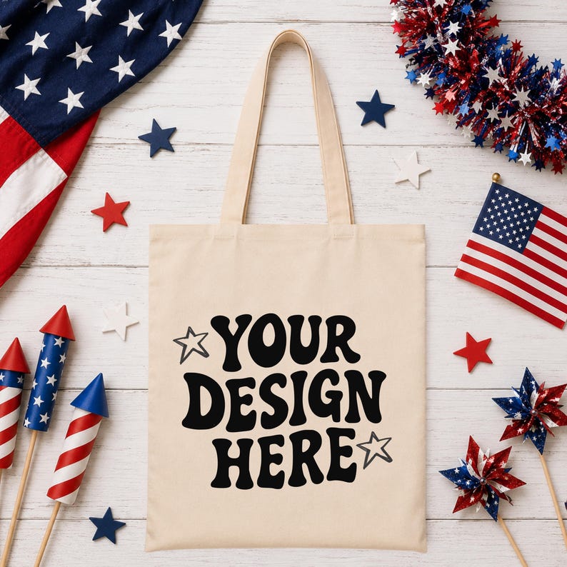 Puede incluir: Un bolso tote color crema con el texto "YOUR DESIGN HERE" en negro. La imagen presenta decoraciones patri&oacute;ticas, incluyendo una peque&ntilde;a bandera americana, adornos en forma de estrella y fuegos artificiales rojos, blancos y azules.
