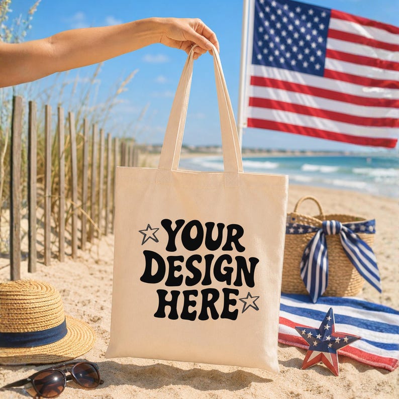 Puede incluir: Un bolso tote beige con el texto "YOUR DESIGN HERE" en negro, sostenido en una playa. El fondo incluye un sombrero de paja, gafas de sol, una cesta con un lazo a rayas azules y blancas y una bandera estadounidense.