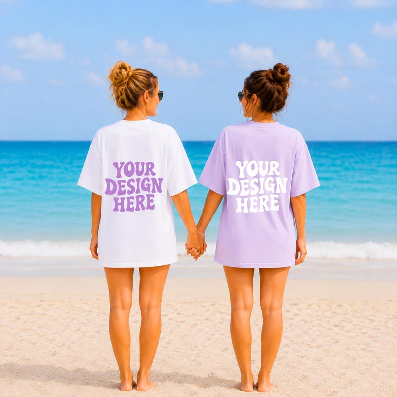 Puede incluir: Dos personas con camisetas blancas y lavanda con el texto "YOUR DESIGN HERE" en una fuente retro. Est&aacute;n de pie en una playa de arena con el oc&eacute;ano de fondo. El cielo es azul y la escena es brillante y soleada.