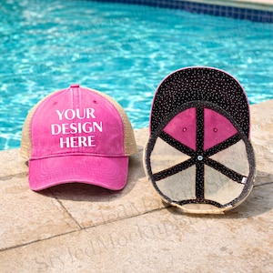 Puede incluir: Dos gorras de camionero rosas con el texto "YOUR DESIGN HERE" en blanco. Una gorra está en posición vertical, la otra está volteada, revelando el diseño interior. Las gorras tienen una parte trasera de malla y están sobre una superficie de piedra.