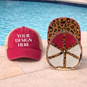 Puede incluir: Gorra de camionero roja con el texto "YOUR DESIGN HERE" en blanco. La gorra tiene una parte trasera de malla y un diseño de estampado de leopardo en la parte inferior de la visera. La gorra se muestra sobre una superficie de piedra.