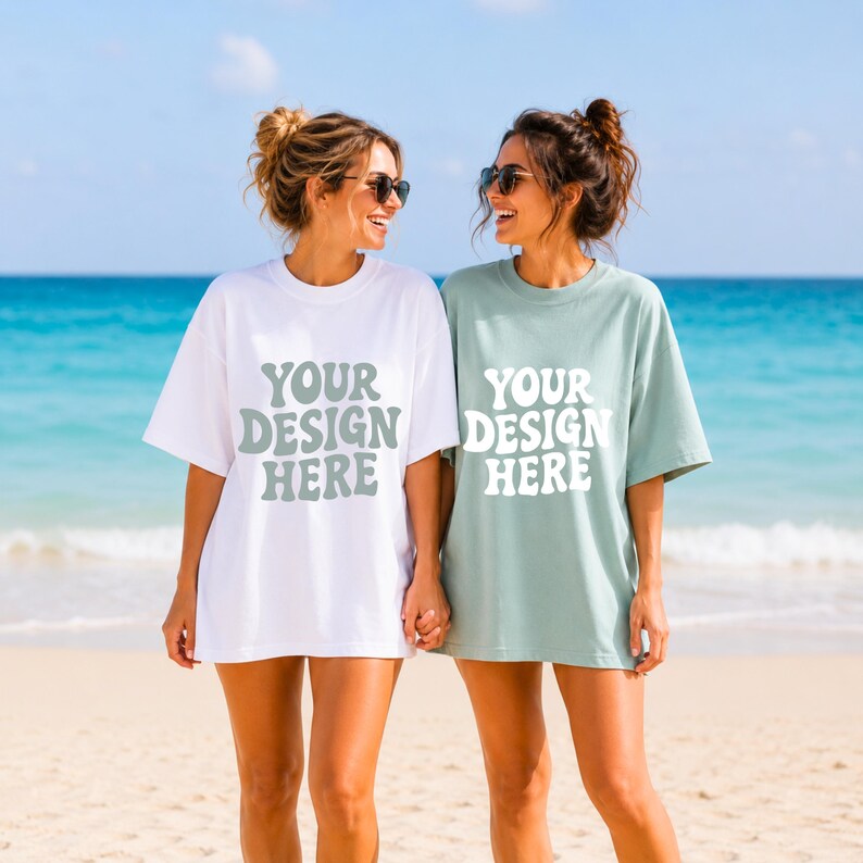 Puede incluir: Dos personas con camisetas de gran tama&ntilde;o en una playa. Una camiseta es blanca, la otra verde apagado. Ambas camisetas tienen el texto "YOUR DESIGN HERE" en una fuente retro. El fondo presenta un oc&eacute;ano y cielo azules.