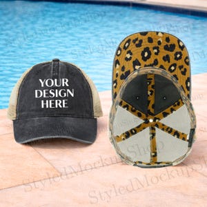 Puede incluir: Tres gorras de béisbol. Una es negra con el texto "YOUR DESIGN HERE" en blanco. La segunda gorra tiene un diseño de estampado de leopardo. La tercera gorra es negra con un diseño de estampado de leopardo en la parte inferior de la visera. Las gorras están sobre una superficie de piedra.