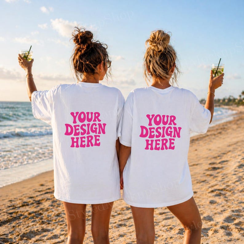 Puede incluir: Dos camisetas blancas con el texto "YOUR DESIGN HERE" en rosa, usadas por dos personas en una playa. Las personas sostienen bebidas con pajitas. El fondo incluye el oc&eacute;ano y una playa de arena.