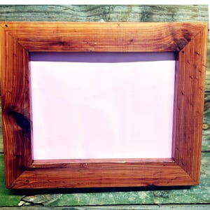 Puede incluir: Un marco de fotos rectangular de madera con un acabado de madera natural. El marco tiene un borde ancho y un interior rosa, perfecto para exhibir una fotografía o una obra de arte. El marco está sobre una superficie de madera verde desgastada.