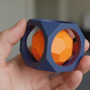 May include: A geometric puzzle toy featuring a navy blue outer frame with hexagonal openings, encasing an orange, multi-faceted sphere. The toy is held in a hand, showcasing its design and color contrast.