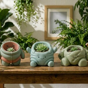 May include: Three whimsical ceramic planters in shades of green and pink, each with a succulent plant. The planters have a cartoonish design with arms and legs, sitting on a wooden table. The background includes greenery and a framed picture.