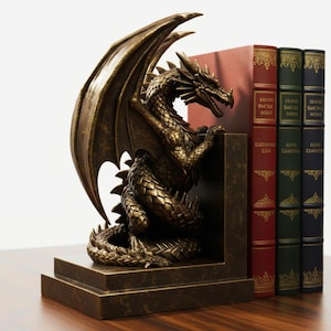 May include: Bronze dragon bookends with detailed wings and scales. The dragon is positioned next to a stack of books with red, green, and blue covers. The bookends are made of a bronze-colored material and sit on a wooden surface.