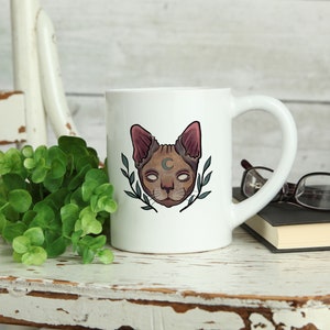 May include: White ceramic mug featuring a stylized cat face with a crescent moon and leafy accents. The cat has a brown face, pink ears, and white eyes. The mug is on a distressed white wooden surface with greenery.
