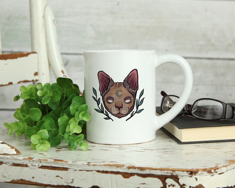 May include: White ceramic mug featuring a stylized cat face with a crescent moon and leafy accents. The cat has a brown face, pink ears, and white eyes. The mug is on a distressed white wooden surface with greenery.