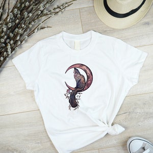 May include: White t-shirt featuring a cat illustration perched on a crescent moon with floral accents. The design incorporates shades of purple, pink, and brown. The shirt is knotted at the waist and paired with gray sneakers and a straw hat.