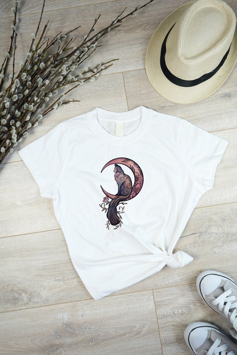 May include: White t-shirt featuring a cat illustration perched on a crescent moon with floral accents. The design incorporates shades of purple, pink, and brown. The shirt is knotted at the waist and paired with gray sneakers and a straw hat.