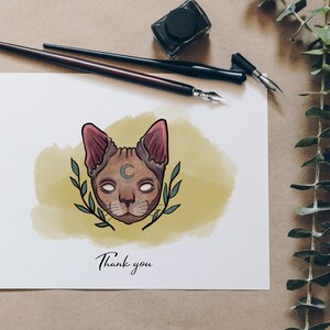 May include: A greeting card with a cat illustration. The cat has a crescent moon on its forehead and is surrounded by leaves. The words "Thank you" are written below the cat. Two calligraphy pens and an ink bottle are on the table.