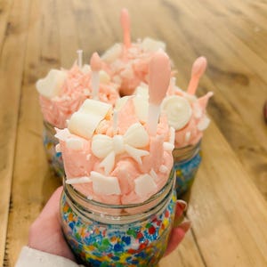 May include: Four decorative candles in glass jars. Each jar is filled with colorful beads and topped with pink frosting, white chocolate pieces, and a bow. The candles have a pink and white color scheme.