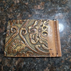 May include: A brown leather money clip with an embossed floral and scroll design in gold and brown tones. The money clip has a lighter brown leather edge.