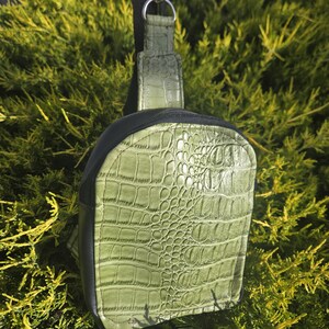 May include: A small, olive green crossbody bag with a crocodile skin texture. The bag has a black zipper and trim, a black strap, and a silver-colored clasp. The bag is shown against a backdrop of green foliage.