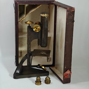 May include: A dark metal microscope with brass adjustment knobs and a stage, positioned in front of an open, worn, dark red storage case. Two separate brass objective lenses are placed on the surface in front of the microscope base.