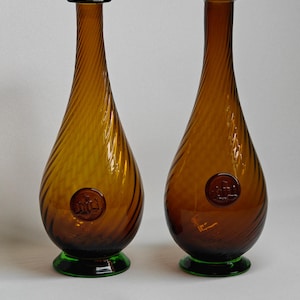 May include: Two amber-colored glass bottles with a spiral design. Each bottle has a dark brown top and a green base. A circular emblem is embossed on the front of each bottle. The bottles are teardrop-shaped and have a vintage aesthetic.