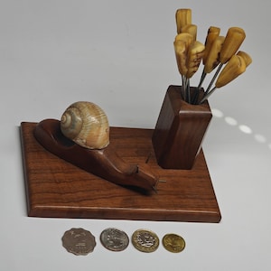 May include: A wooden snail sculpture with a real snail shell on its back, positioned on a rectangular wooden base next to a wooden holder containing several small metal picks with light-colored wooden handles, accompanied by four coins.