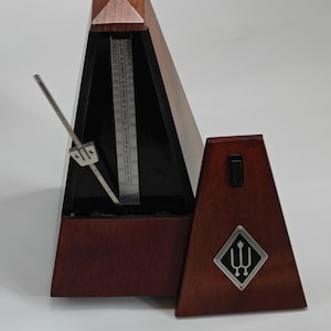 May include: A dark wood mechanical metronome with a pyramid shape, featuring a silver metal pendulum arm and a central scale with tempo markings. A matching wooden cover sits to the right, displaying a silver diamond-shaped logo with a tuning fork.