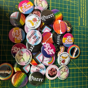 Op de afbeelding: Een verzameling kleurrijke buttons met diverse ontwerpen en tekst. De buttons tonen zinnen als "Queer", "Love is Love" en "Protect the Dogs". Sommige buttons tonen regenboog- en pride vlag ontwerpen.