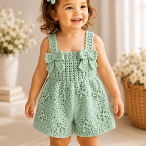 May include: A light sage green crochet romper featuring a square neckline, two large bows on the bodice, and smaller bow accents scattered across the shorts. The garment includes a matching sage green crochet flower hair clip with a white centre.