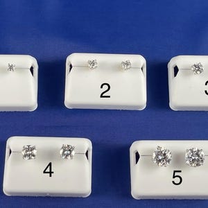 May include: Five pairs of diamond stud earrings displayed on white earring cards against a blue background. Each card is labelled with a number from 1 to 5. The earrings vary in size and cut, showcasing different styles.