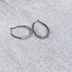 May include: A pair of silver hoop earrings. One earring is in focus, showing a row of small, clear crystals set along the front half of the hoop. The other earring is slightly out of focus, with a similar design. The earrings are on a white, textured surface.